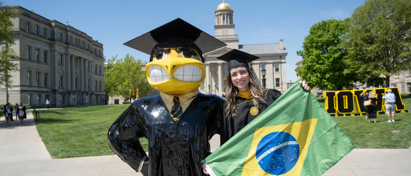 graduating student holding Brazilian flag standing near Herky statue 