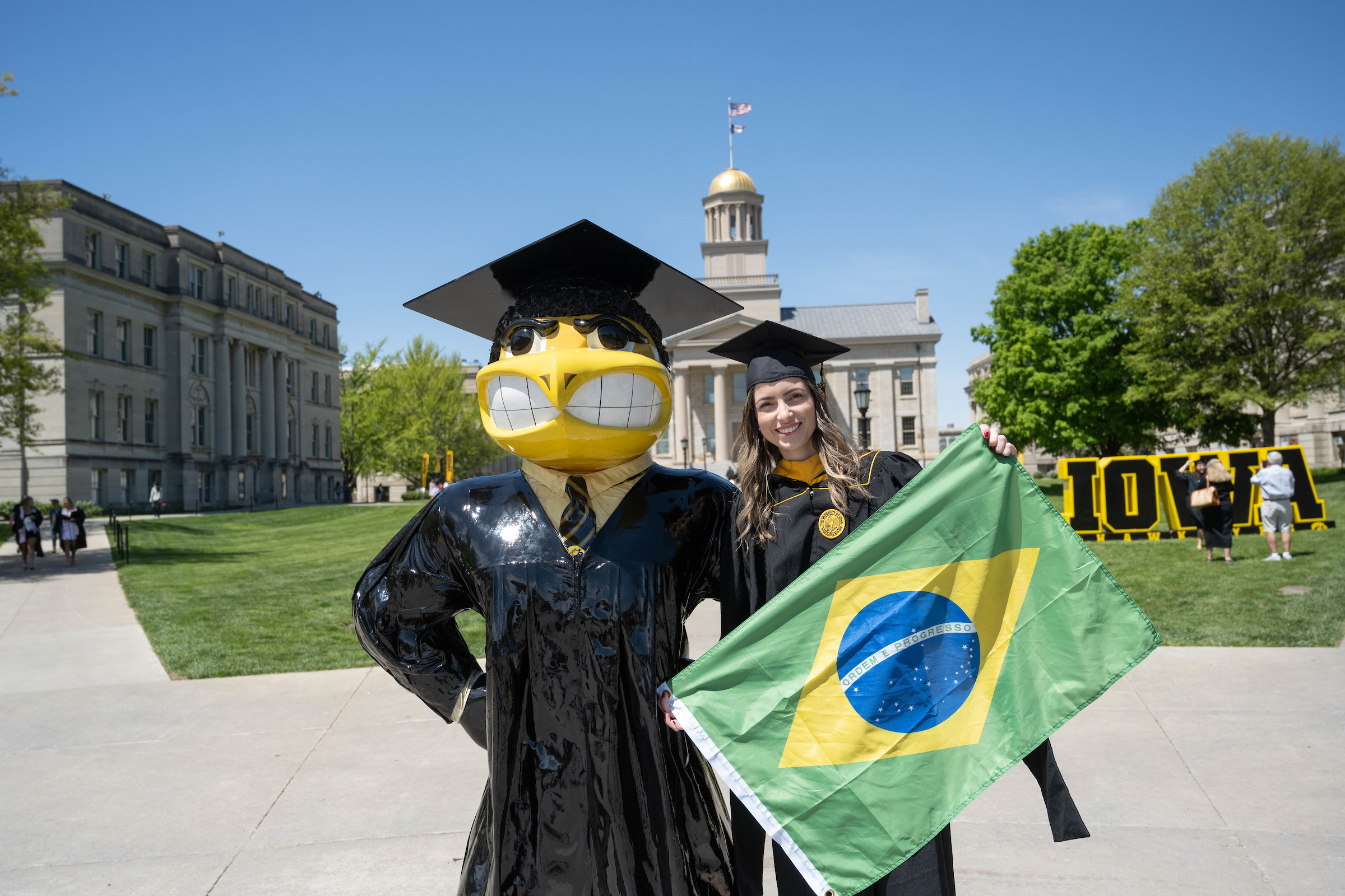 graduating student holding Brazilian flag standing near Herky statue 