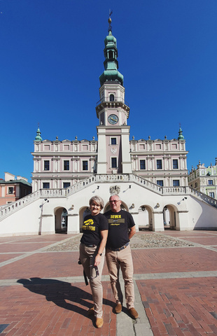 A couple stands together in a large, open plaza with a historic building and wide staircase in the background under a clear blue sky.