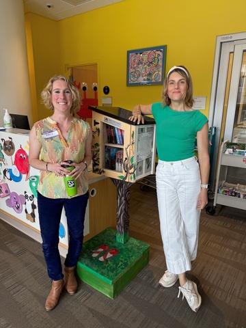 two directors in colorful tops leaning on the Little Free Library box, yellow background