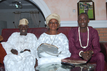 Three people sitting on a couch, dressed in traditional attire. Two wear white, one wears maroon. A handbag is on the table in front of them. A framed picture hangs on the wall behind.