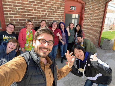 A group of ten smiling people wave in front of a brick building, enjoying a sunny day. The atmosphere is joyful and casual.