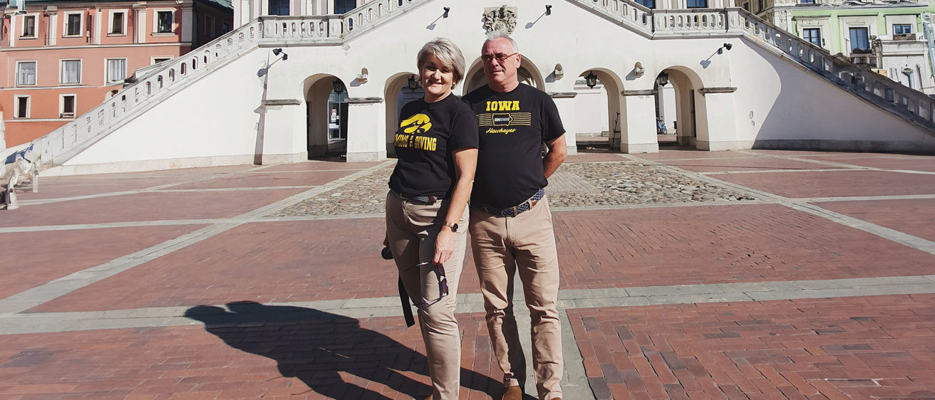 A couple stands together in a large, open plaza with a historic building and wide staircase in the background under a clear blue sky.