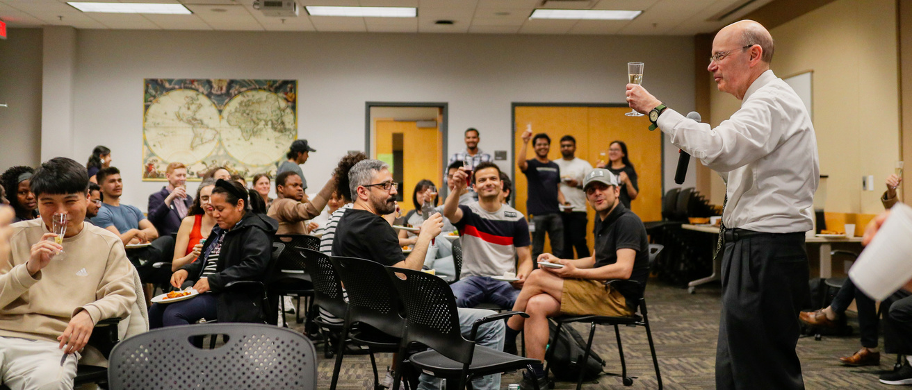a crowd of student sitting and raising a glass as Russ Ganim gives a toast to international students completing the academic year.