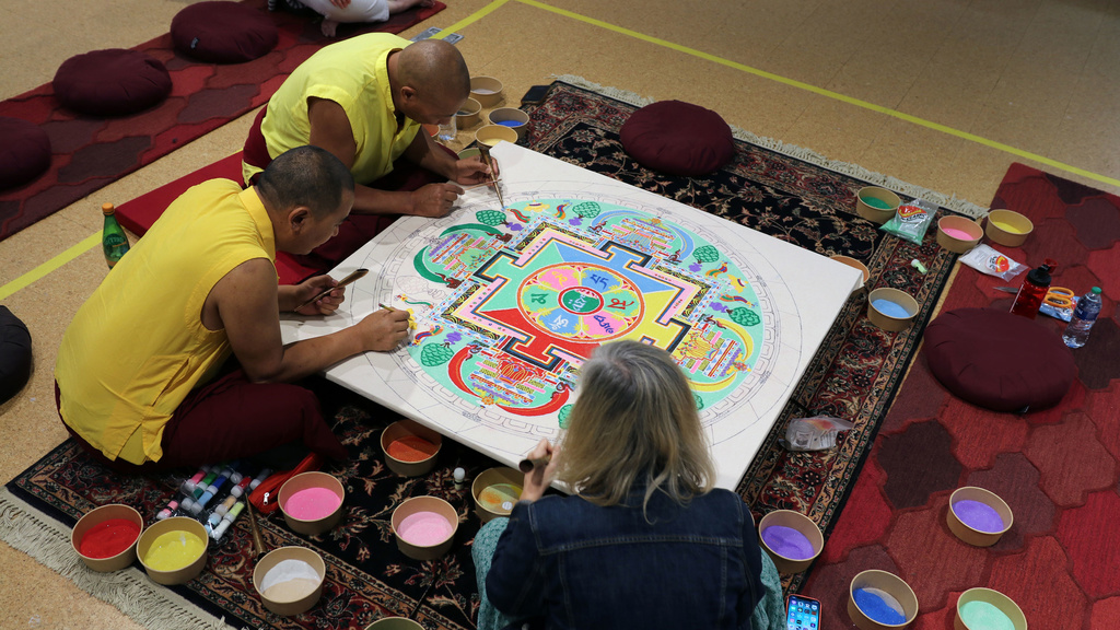 Two men in Buddhist yellow shirts and woman work on colorful sand mandala