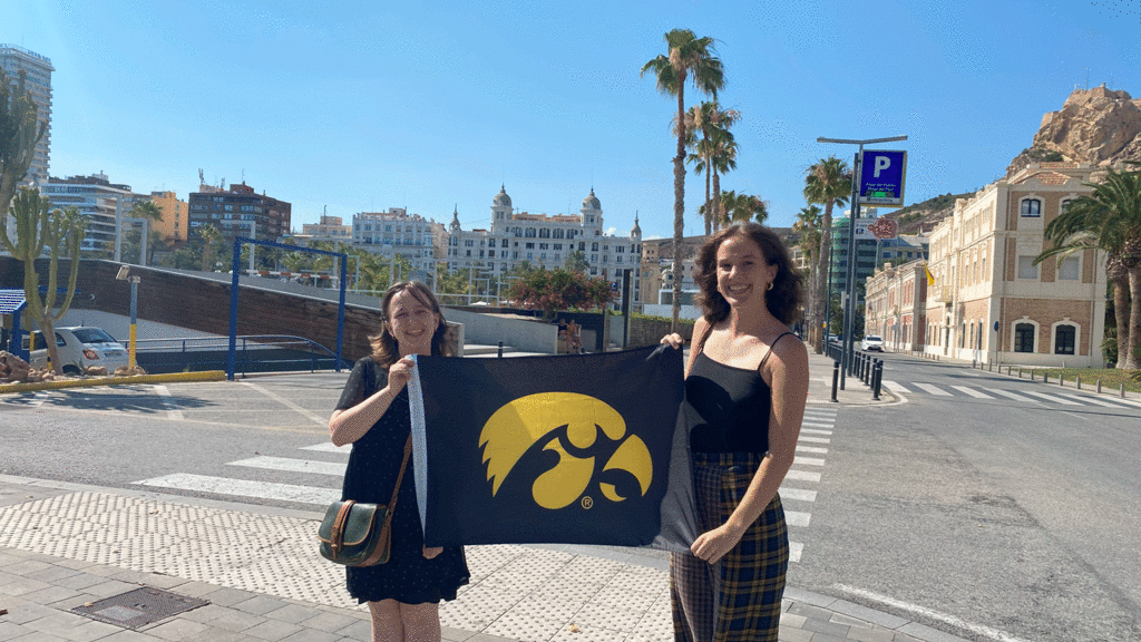 UI students MK Shultz and Megan Slinger in Alicante, Spain holding Hawkeye flag in front of palm tree lined streets of Alicante